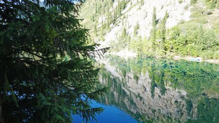 Kolsay lake located in the mountains of Kazakhstan. Turquoise water as a mirror. The water reflects the mountains, green hills, tall spruce, grass, sky and clouds. Beautiful landscape of mountain lake.の写真素材