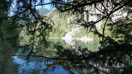 Kolsay lake located in the mountains of Kazakhstan. Turquoise water as a mirror. The water reflects the mountains, green hills, tall spruce, grass, sky and clouds. Beautiful landscape of mountain lake.の写真素材