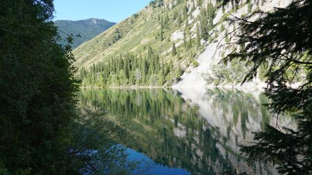 Kolsay lake located in the mountains of Kazakhstan. Turquoise water as a mirror. The water reflects the mountains, green hills, tall spruce, grass, sky and clouds. Beautiful landscape of mountain lake.の写真素材