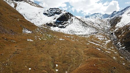 Autumn in the mountains. Yellow grass and snow-capped mountains. The gorge with the river. Blue sky and big white clouds. The stones lie on the slopes, in some places the snow. Almaty, Kazakhstan.の写真素材