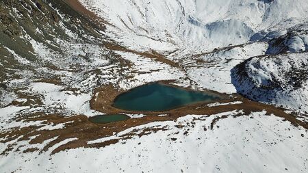 Mountain lake among the snowy peaks. Moraine lake Titova, in the mountains of Almaty, Kazakhstan. Height 3377 m. Shooting with a drone. Yellow rocks, white clouds, snow and blue sky. Highest peak.の写真素材