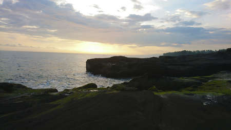 Sunset over the ocean. View of large waves breaking on the rocks. Clouds hang over the horizon. A ray from the sun is reflected on the water in orange color. The coast of Bali is visible.の写真素材