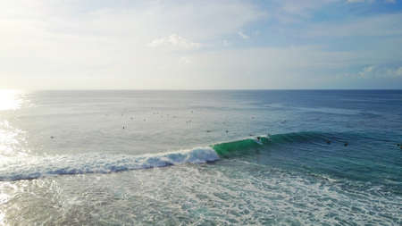 Surfers catch waves at Uluwatu beach, Bali. Huge waves stretch along the beach. A lot of people with boards are catching waves. The wave swirls into the tube, a huge foam. View from a drone.の写真素材
