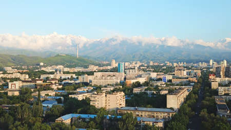 Bright color sunset over the city of Almaty. Huge clouds over the mountains and the city shimmer from bright blue to yellow and dark blue. Tall houses and green trees, cars driving on the roads.の写真素材