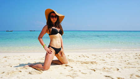 The girl in the hat and swimsuit on the beach. Glasses on. European woman on the beach with white sand. The water is turquoise with a blue tint. Photo shoot model. White sand and blue sky. Paradise.の写真素材