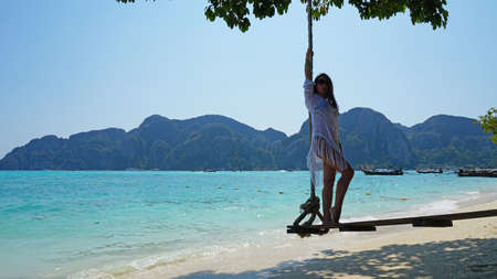 Beautiful girl in a white Cape and a black bathing suit on a swing. Swing on the beach. View of the island, beach with sand, blue water and green leaves of trees. Visible hills of the island. Thailandの写真素材