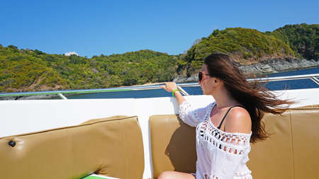Girl sitting on the nose of the speedboat. Hair fluttering in the wind, glasses in front of his eyes. White robe. View of green island and blue sea. Shoulders open. Comfortable leather seats.の写真素材