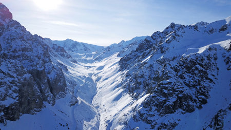 High snowy mountains, spruce trees grow in places. Huge rocks and cliffs, gorges where there may be an avalanche. Top view from a drone. Sunny day, shadows from the mountains fall on the snow. Almatyの写真素材