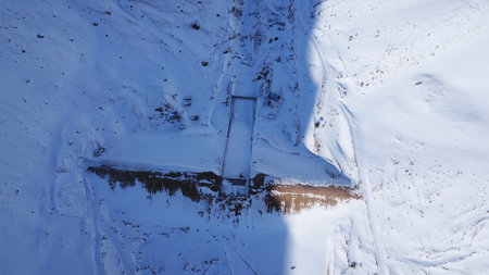 The dam was covered with snow in the mountains. The view from the top. The river is frozen, everything is covered with snow. Equipment, houses, a gorge in the snow. Shadow from the sun. Mountain dam.の写真素材