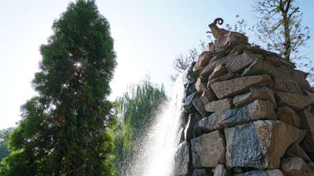 Artificial waterfall with a statue in the city Park of Almaty. Mountain sheep stands on the high stones of the waterfall, water flows into the bowl. Green trees and sky all around.の写真素材