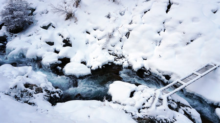 A clear mountain river runs through a snowy gorge. There is a metal bridge covered with snow. Tall spruce trees grow on the slopes of the mountains. There is steam from the river. Almarasan, Almatyの写真素材