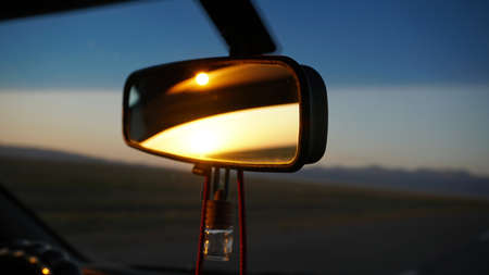 The rear view mirror of the car and dawn. Orange dawn over the hills. The car is going at high speed. Green fields, grass and meadows are visible. Black color of the car. Steering wheel, hood, glass.の写真素材