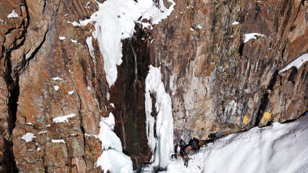 Freezing waterfall in the snowy mountains. View from the drone, from above. The rocks are covered with snow and ice. A small stream of water runs. The waterfall freezes. A group of people are restingの写真素材