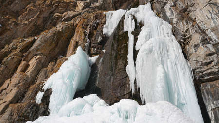 Frozen waterfall among the rocks. The waterfall is freezing, huge icicles. Ice white and blue. Brown rocks and splashes of water. Winter waterfall. White snow and blue sky. Water runs down the ice.の写真素材
