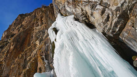Frozen waterfall among the rocks. The waterfall is freezing, huge icicles. Ice white and blue. Brown rocks and splashes of water. Winter waterfall. White snow and blue sky. Water runs down the ice.の写真素材