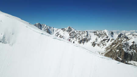 A group of climbers climb to the top of the peak. Dangerous, steep slope. Avalanche-prone area. Mountains covered with snow. Steep cliffs and large rocks. Above the mountains, top view from a drone.の写真素材