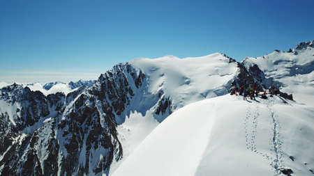 A group of climbers climb to the top of the peak. Dangerous, steep slope. Avalanche-prone area. Mountains covered with snow. Steep cliffs and large rocks. Above the mountains, top view from a drone.の写真素材