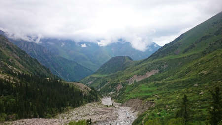 The river runs through a green gorge in the mountains. The view from the height of the bustling river, large stones, green fields and trees. The hills are covered with clouds. Ala Archa, Kyrgyzstan.の写真素材
