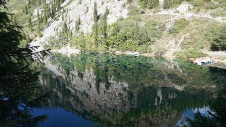 Kolsay lake among green hills and mountains. The mountain lake is surrounded by green forest, tall coniferous trees, grass and bushes. Clean water is like a mirror. Tourists swim on boats. Kazakhstanの写真素材