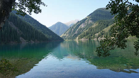 Kolsay lake among green hills and mountains. The mountain lake is surrounded by green forest, tall coniferous trees, grass and bushes. Clean water is like a mirror. Tourists swim on boats. Kazakhstanの写真素材