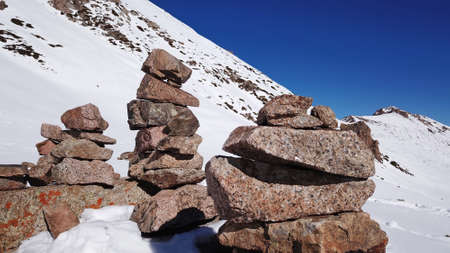 Stone pyramid pointer tour in the snowy mountains. Stone pyramids serve as pointers to the route. All around is snow, peaks, blue sky and rocks. Climbing in the mountains. Almaty, Kazakhstan.の写真素材