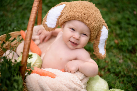 Cute little baby boy in suit of rabbit sitting on the grass in basket with cabbage and carrot. Nature park backgroundの写真素材