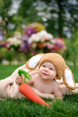 Baby in the hat like a bunny with carrot toy on green grass outdoorsの写真素材