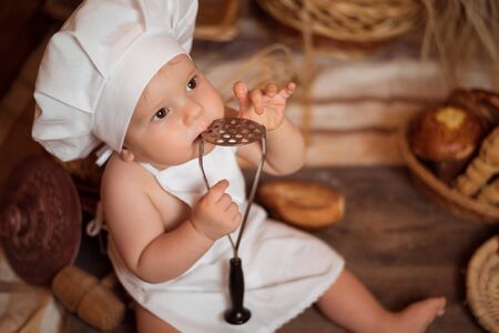Happy little child in the cook costume at the kitchen sitting on the table among baskets with tasty rolls, buns and bread. Copy spaceの写真素材