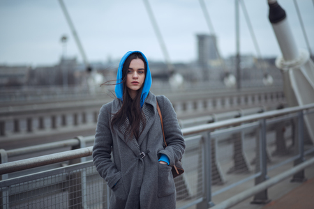Young woman in the coat, walks on the city bridge during the windy weather. Urban autumn winter styleの写真素材