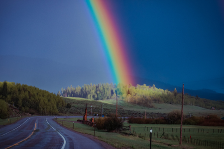 A rainbow ends and brightens a group of trees on a country hillside that surrounds the road.の写真素材