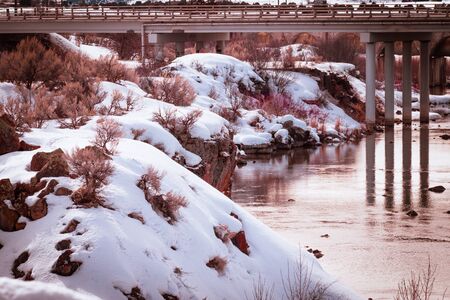 Sun sets and creates colorful reflections in the still water below the bridge at the Bear River Dam. Snow and drided sagebrush cover the rocky cliffs surrounding the low-lying river below.の写真素材