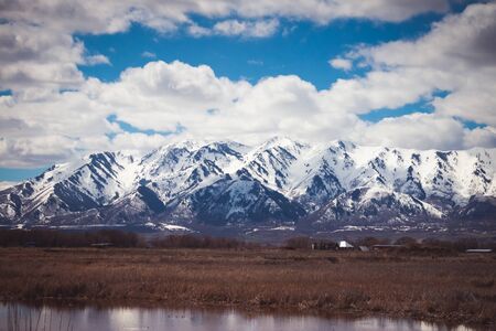 A snow-covered mountain in Logan, Utah sits above a drab Spring farm and field, with a pond reflecting in the foreground.の写真素材
