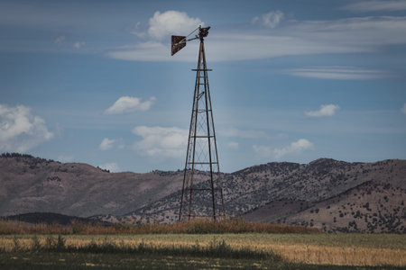 The remains of an old broken windmill sits abandoned in a rural field, against a bright blue sky and desert mountains.の写真素材
