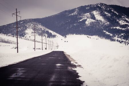A road to nowhere ends at a scenic winter view of the mountainside and snow-covered field. The road is wet with puddles and bordered by electric poles.の写真素材