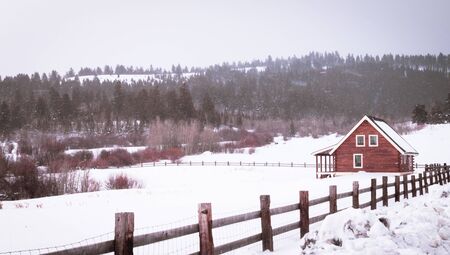 A little red farm cabin sits in a snow-covered field, surrounded by mountain scenery.の写真素材