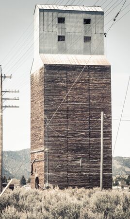An old grain elevator in Soda Springs, ID sits abandoned next to railroad tracks, surrounded by overgrown landscape, electrical wires and a junkyard of rusty vintage farm equipment and machinery parts.の写真素材