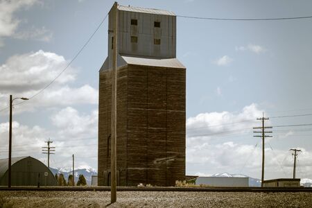 A vintage grain elevator in Soda Springs, ID sits abandoned next to railroad tracks, surrounded by electrical wires, overgrown landscape and a junkyard of rusty old farm equipment and machinery parts.の写真素材