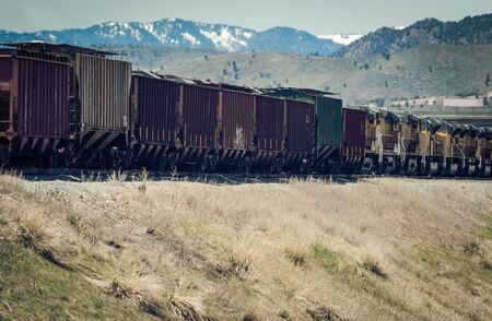 Classic yellow Union Pacific diesel locomotive engines transport rusty retro grain hopper train cars next to a grassy ravine in a rural country town with snowy mountains in the background.の写真素材