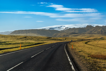 A winding road stretches towards towering mountains and glaciers in Iceland, with a dramatic sky overheadの写真素材