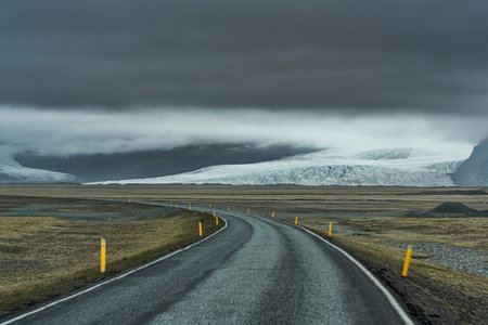 A winding road stretches towards towering mountains and glaciers in Iceland, with a dramatic sky overheadの写真素材