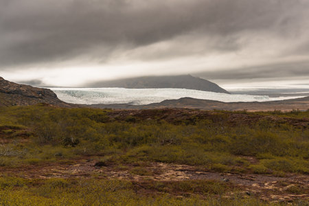A winding road stretches towards towering mountains and glaciers in Iceland, with a dramatic sky overheadの写真素材