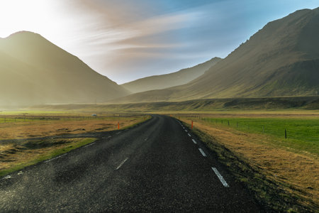 A solitary road winds through the majestic Icelandic mountains, illuminated by the warm hues of a stunning sunset. This serene scene captures the tranquil beauty and rugged charm of Iceland's remote wildernessの写真素材