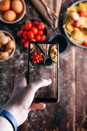 hands picking up a smartphone taking a picture of a fresh pasta on wooden table with ingredients....