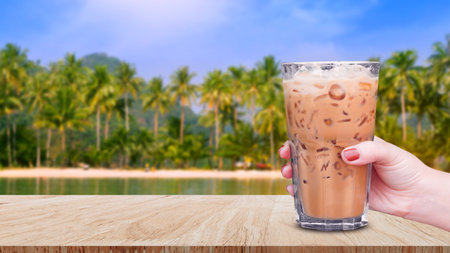 Hand holding the glass iced coffee on wooden table with beach landscape view nature background, Ice latte coffeeの写真素材