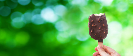 Chocolate covered ice cream bar on Green bokeh background, Hand holds delicious chocolate and nuts almond ice cream bar on a stick. in the summerの写真素材