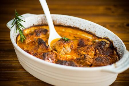 baked meat with spices vegetables and gravy in a ceramic form, on a wooden tableの写真素材