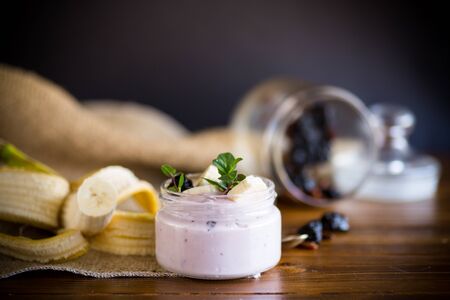 sweet tasty yogurt with banana, prunes, raisins in a glass jar on a wooden tableの写真素材