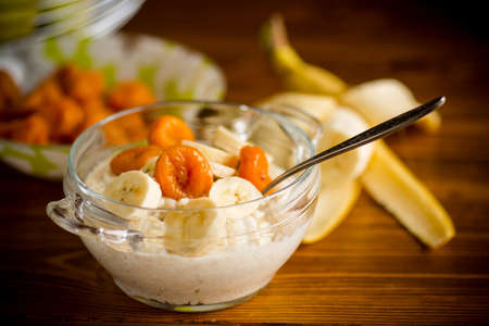 cooked boiled sweet oatmeal with with dried apricots and bananas in a bowl on a wooden tableの写真素材