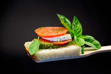 fresh slice of bread with pesto sauce, mozzarella and tomato on a wooden spatula with basil. Isolated on a black background.の写真素材