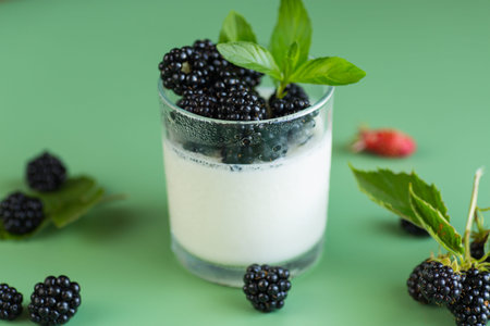 Ripe blackberries with homemade yogurt in a glass on a green background.